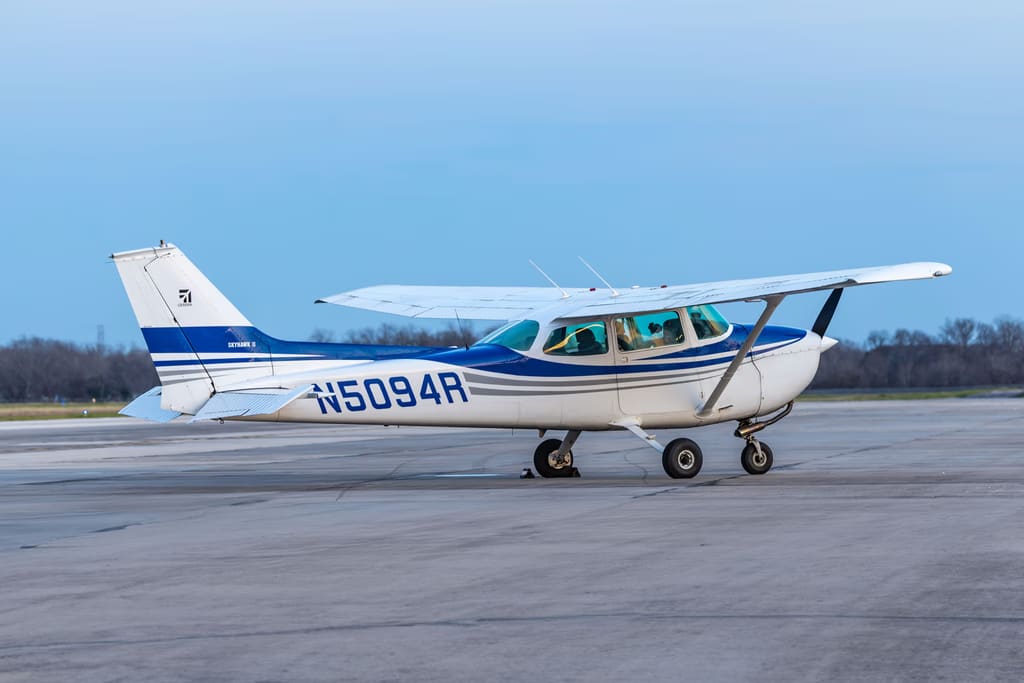 Cessna 172M N5094R in white with blue and gray striping, side-on at the taxiway edge under a clear sky.