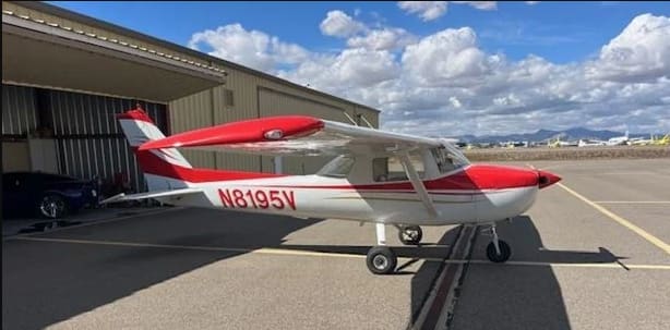 Cessna A150M Aerobat N8195V in white with red trim, parked on the apron beside an open hangar.